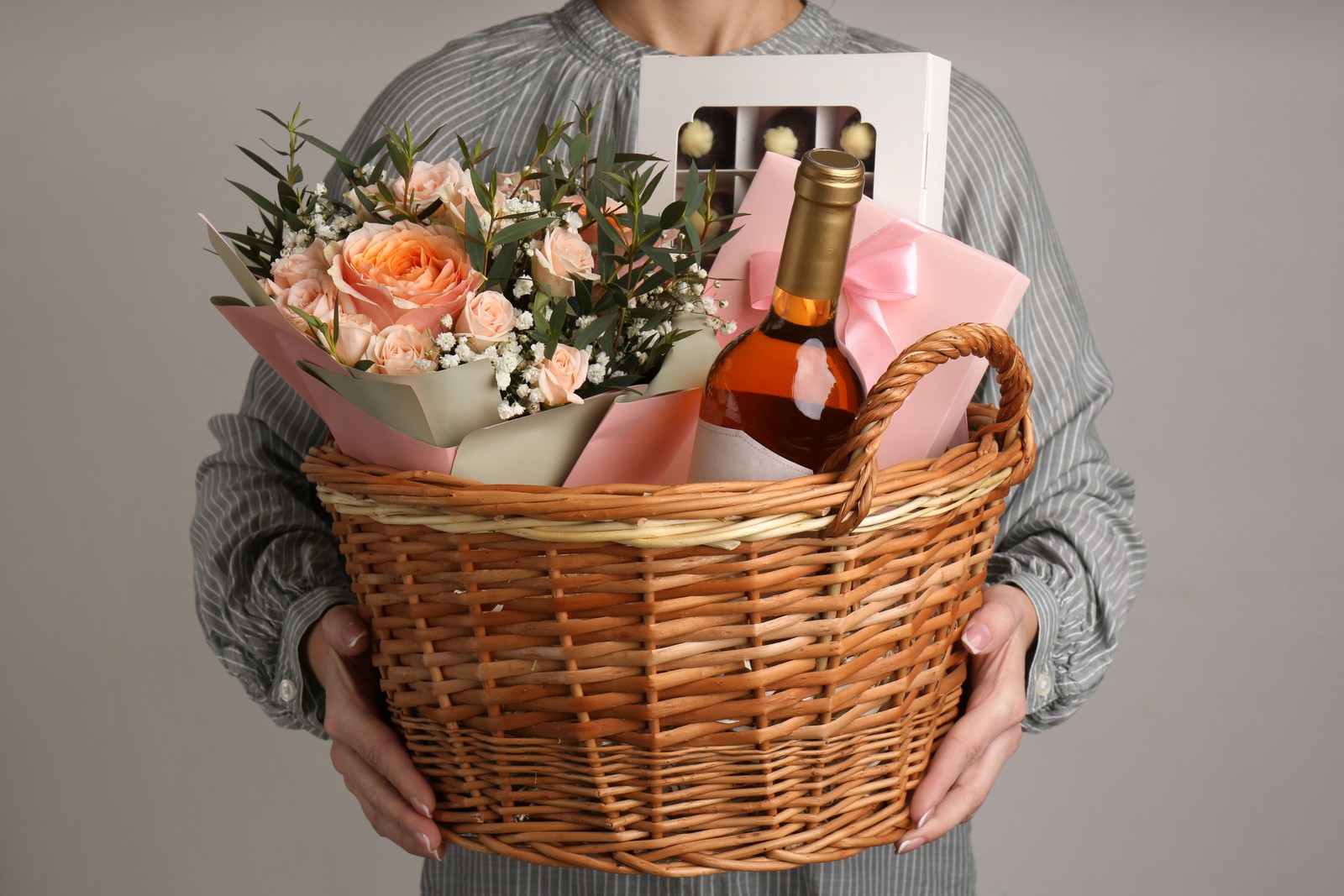 Photo of woman holding wicker basket with different gifts on grey background, closeup