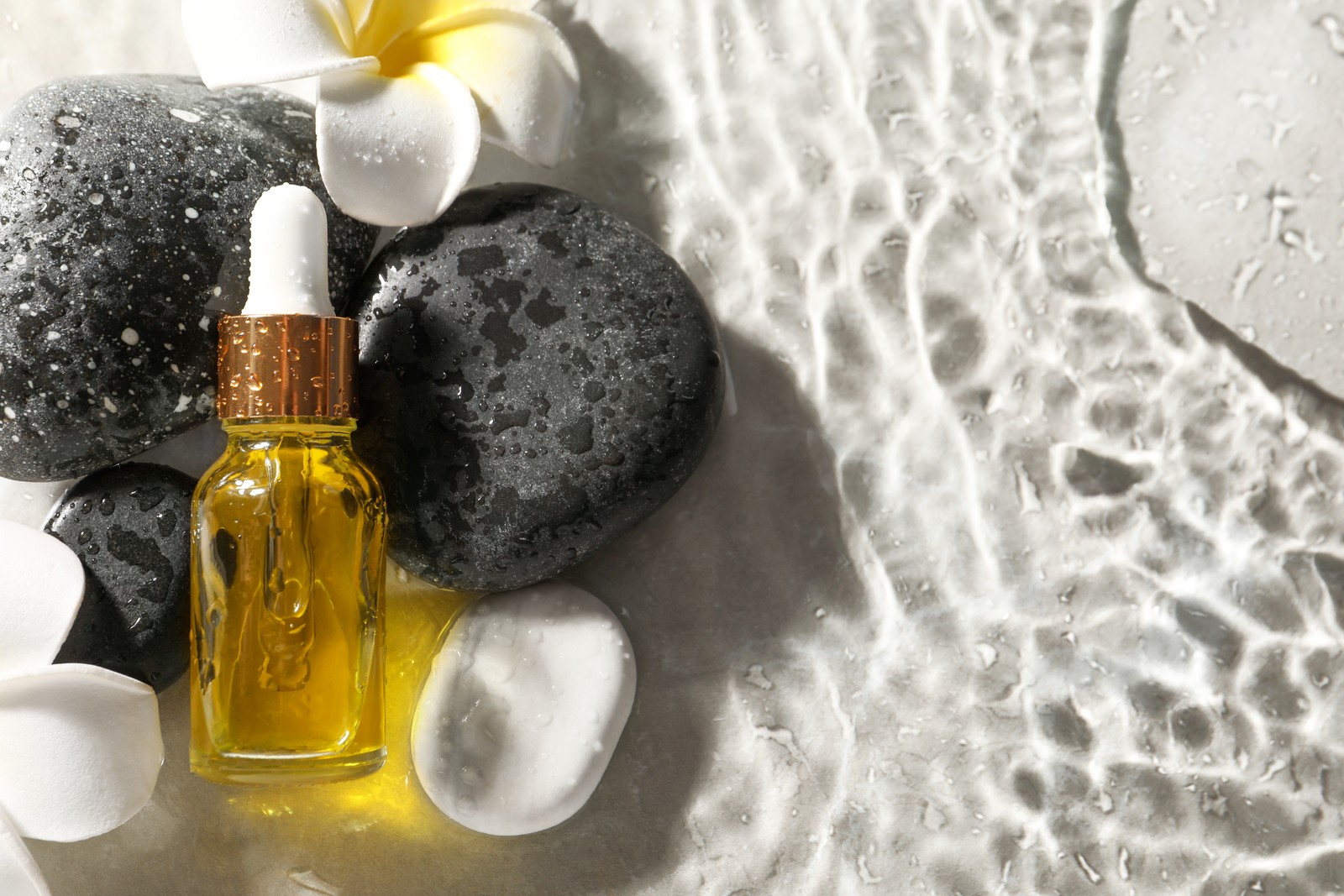 Photo of bottle of face serum and spa stones in water on light background, flat lay. Space for text