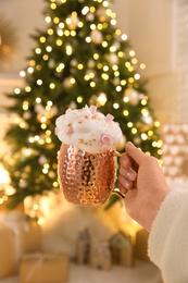 Woman holding cup of delicious drink with whipped cream and marshmallows near Christmas tree indoors, closeup