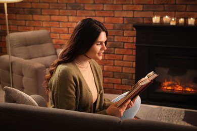 Young woman reading book on sofa near fireplace at home
