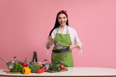 Young housewife with pan, spatula and products on pink background