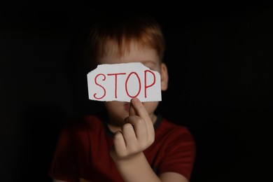 Photo of Little boy holding piece of paper with word Stop in darkness, focus on hand. Domestic violence concept