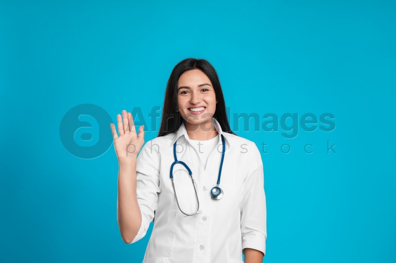 Happy female doctor waving to say hello on light blue background