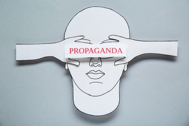 Photo of Information warfare. Human closing eyes with hands and card with word Propaganda. Paper cutouts on light grey background, top view