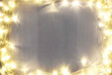 Frame of beautiful glowing Christmas lights on white wooden table, top view. Space for text