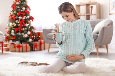 Happy pregnant woman with cup of tea sitting on floor in room decorated for Christmas