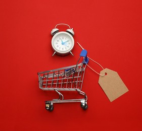 Shopping cart with tag and alarm clock on red background, flat lay. Sale concept