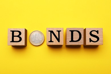 Photo of Word Bonds made of wooden cubes with letters and coin on yellow background, flat lay