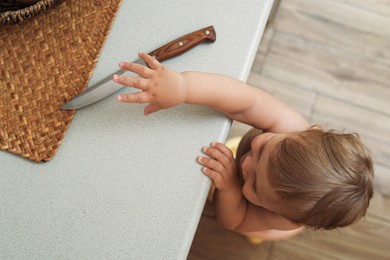 Photo of Little child reaching for knife on light countertop, above view. Dangers in kitchen