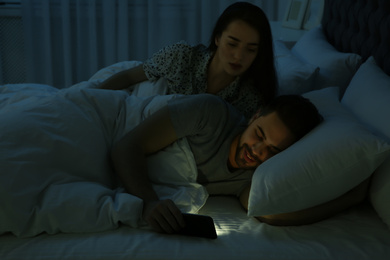 Distrustful young woman peering into boyfriend's smartphone in bed at night