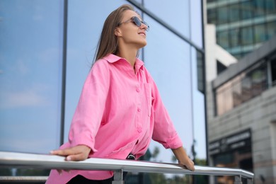 Beautiful young woman in stylish sunglasses holding onto railing near building outdoors