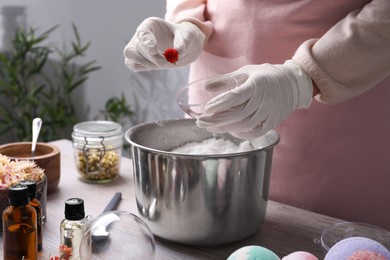 Woman in gloves putting dried flower into bath bomb mold at wooden table indoors, closeup