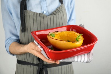 Photo of Woman holding baking dish with half of cooked spaghetti squash and basil on light background, closeup