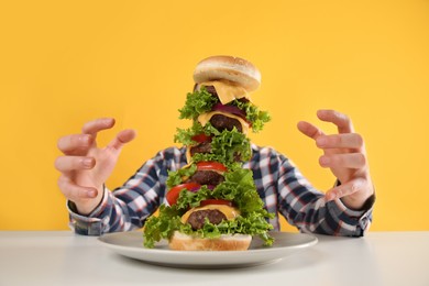 Hungry man with huge burger at white table on yellow background
