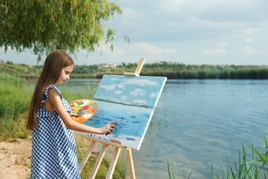 Photo of Little girl painting scenery on easel near lake