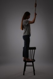Depressed woman with rope noose standing on chair against grey background