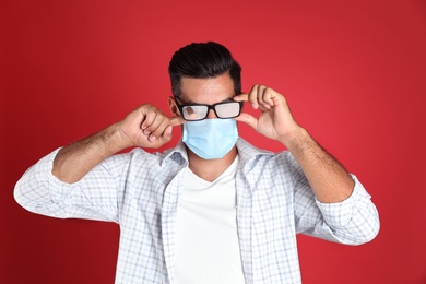 Man wiping foggy glasses caused by wearing medical mask on red background