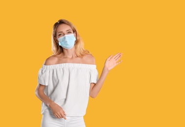 Woman in protective face mask showing hello gesture on yellow background, space for text. Keeping social distance during coronavirus pandemic