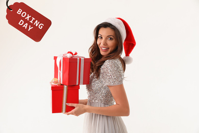 Beautiful woman wearing Santa hat holding Christmas gifts and tag with text Boxing day on white background