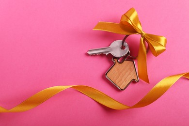 Photo of Key with trinket in shape of house, bow and ribbon on pink background, top view. Space for text. Housewarming party