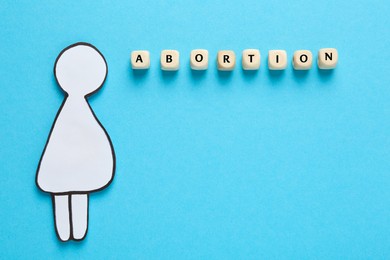 Word Abortion made of wooden cubes and pregnant woman paper cutout on light blue background, flat lay