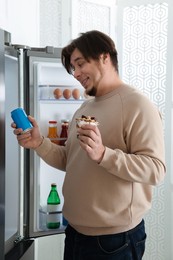 Overweight man holding dessert and tin can with beverage near open refrigerator in kitchen
