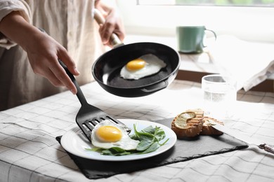 Woman putting tasty fried eggs onto plate at table indoors, closeup