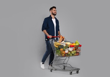 Happy man with shopping cart full of groceries on light grey background
