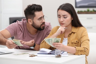 Worried young couple counting money at white table indoors
