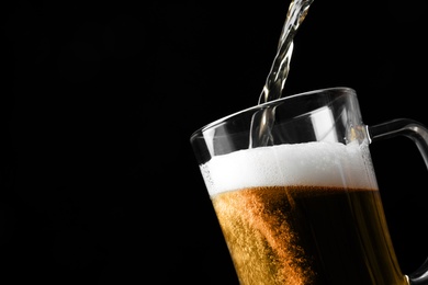 Pouring cold tasty beer into glass mug on black background, closeup. Space for text