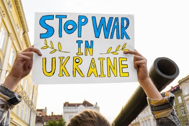 Woman holding poster in colors of national flag with words Stop War In Ukraine near broken military tank on city street, closeup
