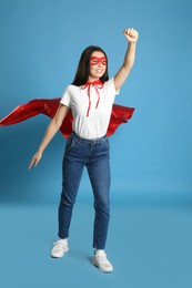 Confident young woman wearing superhero cape and mask on light blue background