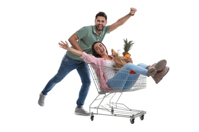 Young man giving his girlfriend ride in shopping cart with groceries on white background