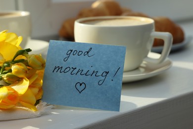 Cup of coffee, flowers and card with phrase GOOD MORNING! on white wooden table, closeup