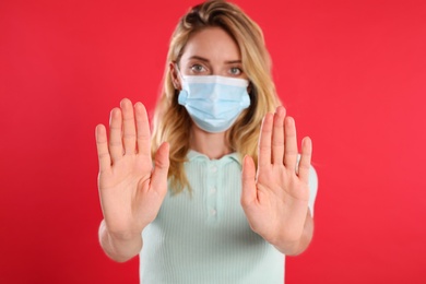 Woman in protective face mask showing stop gesture on red background, focus on hands. Prevent spreading of coronavirus