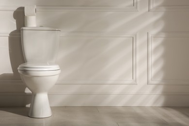Modern toilet bowl and paper rolls near white wall in restroom, space for text. Interior design