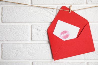 Red envelope and card with lip print hanging on twine near white brick wall, space for text. Love letter