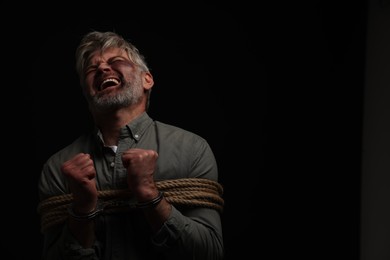 Photo of Emotional beaten man tied with rope on black background, space for text. Hostage taking