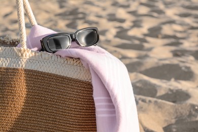 Stylish wicker bag, sunglasses and blanket on sand, closeup