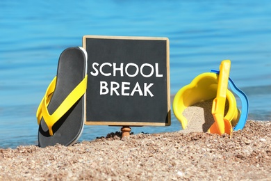 Image of Small black chalkboard with text School Break, beach toys and flip flops on sand near sea. Seasonal holidays 