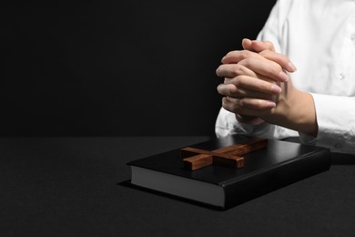 Woman holding hands clasped while praying at table with Bible against black background, closeup. Space for text