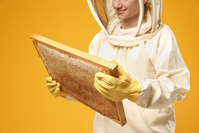 Beekeeper in uniform holding hive frame with honeycomb on yellow background, closeup