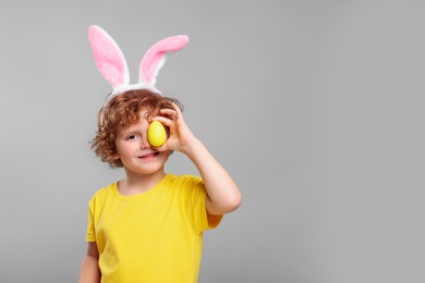 Photo of Happy boy in cute bunny ears headband covering eye with Easter egg on light grey background. Space for text