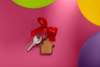 Photo of Key with trinket in shape of house and red bow on pink background near color balloons, flat lay. Housewarming party