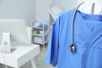 Photo of Blue medical uniform and stethoscope hanging on rack in clinic, closeup. Space for text