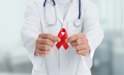 Doctor holding red awareness ribbon on blurred background, closeup. World AIDS disease day