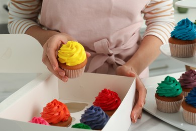Woman with box of delicious colorful cupcakes at white table indoors, closeup