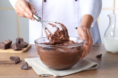 Professional confectioner whipping chocolate cream with balloon whisk at wooden table, closeup