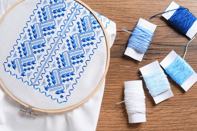 Photo of Shirt with blue embroidery design in hoop and threads on wooden table, flat lay. National Ukrainian clothes