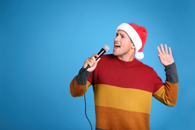 Emotional man in Santa Claus hat singing with microphone on blue background, space for text. Christmas music